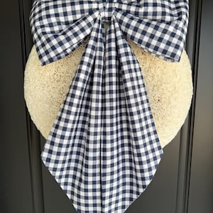 May include: A large decorative bow made of navy blue and white gingham fabric, featuring two wide loops and two long, pointed tails. The bow is centered over a round, textured, off-white loofah-like wreath base, creating a layered visual effect.