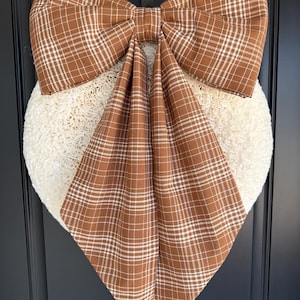 May include: A heart-shaped wreath made of textured, cream-colored fibrous material, adorned with a large, prominent bow made of brown fabric featuring a white plaid pattern. The bow has two wide, long tails that drape downward over the center of the wreath.