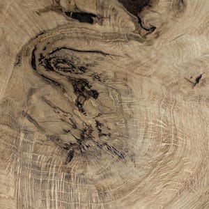Gros plan sur une section de grain de bois, présentant un motif naturel de lignes tourbillonnantes et de marques sombres et irrégulières. La couleur du bois est un brun clair chaud avec des variations de tons, soulignant la texture et les détails organiques.