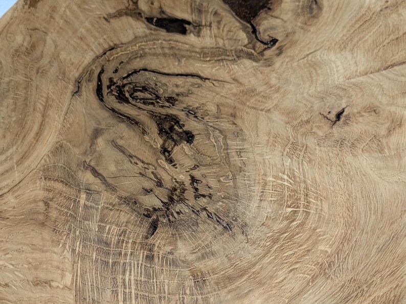 Gros plan sur une section de grain de bois, présentant un motif naturel de lignes tourbillonnantes et de marques sombres et irrégulières. La couleur du bois est un brun clair chaud avec des variations de tons, soulignant la texture et les détails organiques.