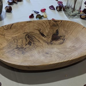 Un plateau en bois de forme ovale avec un motif de grain de bois naturel. Le plateau est brun clair avec des accents brun foncé et une finition lisse et polie. Il est vide et repose sur une surface de couleur claire.
