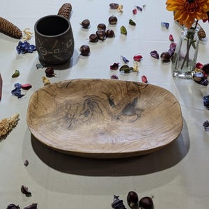 Un plat de service ovale en bois avec un motif de grain naturel. Une tasse en céramique foncée avec des motifs géométriques est à gauche. La scène est décorée de fleurs, de pétales et de châtaignes.