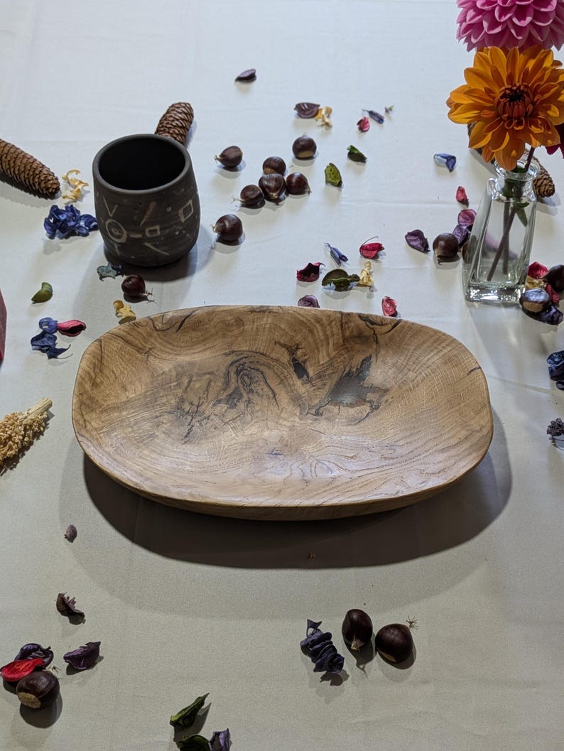 Un plat de service ovale en bois avec un motif de grain naturel. Une tasse en céramique foncée avec des motifs géométriques est à gauche. La scène est décorée de fleurs, de pétales et de châtaignes.