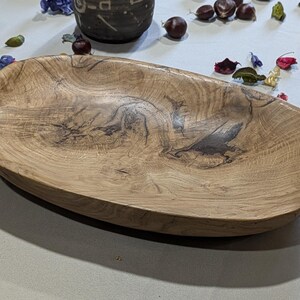 Un plat ovale en bois avec un grain naturel. Le plat est de couleur brun clair avec des motifs plus foncés et tourbillonnants et une finition lisse. C'est une pièce décorative, adaptée au service ou à l'exposition. Mesure environ 30 cm de long.