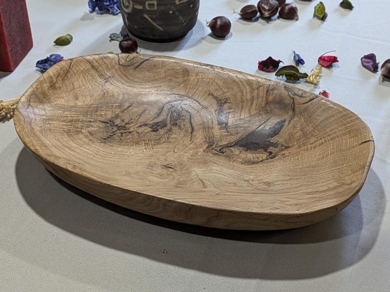 Un plat ovale en bois avec un grain naturel. Le plat est de couleur brun clair avec des motifs plus foncés et tourbillonnants et une finition lisse. C'est une pièce décorative, adaptée au service ou à l'exposition. Mesure environ 30 cm de long.