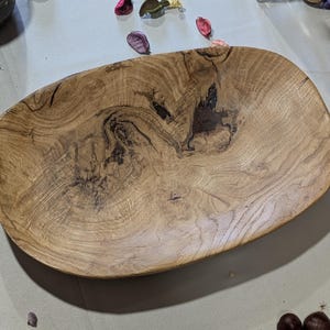 Un plat de service en bois fait à la main, de forme naturelle et organique. Le bol est fait de bois brun clair avec des motifs de grain brun foncé et noir. Le bol ovale est idéal pour servir des aliments ou comme pièce décorative.