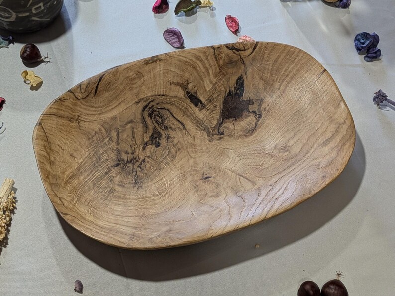Un plat de service en bois fait à la main, de forme naturelle et organique. Le bol est fait de bois brun clair avec des motifs de grain brun foncé et noir. Le bol ovale est idéal pour servir des aliments ou comme pièce décorative.