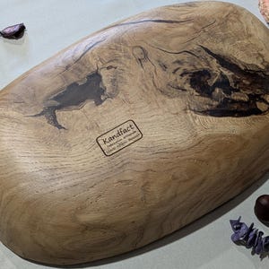 le dessous d'un plateau en bois ovale fait à la main avec une finition naturelle veinée. Le plateau présente des motifs sombres et complexes et une surface lisse et polie. L'étiquette sous le plateau indique "Kandfact". Ce plateau décoratif est parfait pour servir ou exposer.