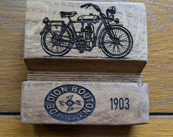 May include: Wooden block featuring a detailed engraving of a vintage motorcycle. The design includes the text "DE DION BOUTON PUTEAUX 1903" in a circular pattern. The motorcycle has a classic design with spoke wheels.