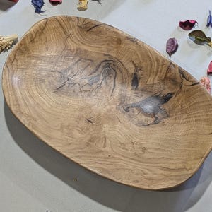 Un plateau ovale en bois avec un motif de grain naturel. Le plat est brun clair avec des stries et des nœuds plus foncés. Des fleurs et des pétales séchés entourent le plat. Idéal pour servir ou décorer.