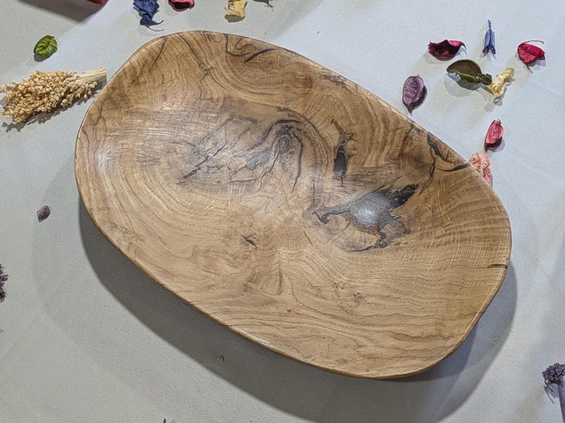 Un plateau ovale en bois avec un motif de grain naturel. Le plat est brun clair avec des stries et des nœuds plus foncés. Des fleurs et des pétales séchés entourent le plat. Idéal pour servir ou décorer.