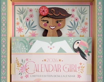 May include: A decorative desk calendar titled "Calendar Girl" for the year 2026, featuring a wooden cutout of a woman with a flower in her hair. The calendar has a tropical theme with toucans and floral designs, presented in a decorative box.