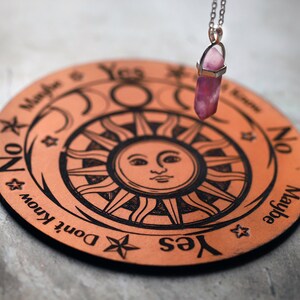 May include: A circular, copper-toned divination board featuring a sun and moon motif, with the words "Yes," "No," "Maybe," and "Don't Know." A pink crystal pendulum hangs above the board, suspended by a silver chain. The board is approximately 15 cm in diameter.