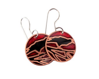 May include: A pair of round earrings with a red and black design. The earrings have a copper-colored border and silver-colored ear wires. The design features a red background with black abstract shapes.