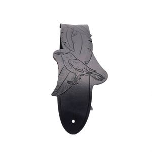 May include: A black guitar strap with a detailed raven and feather design. The strap features a gradient effect, transitioning from light gray to black. A single hole is visible at the bottom for attachment.