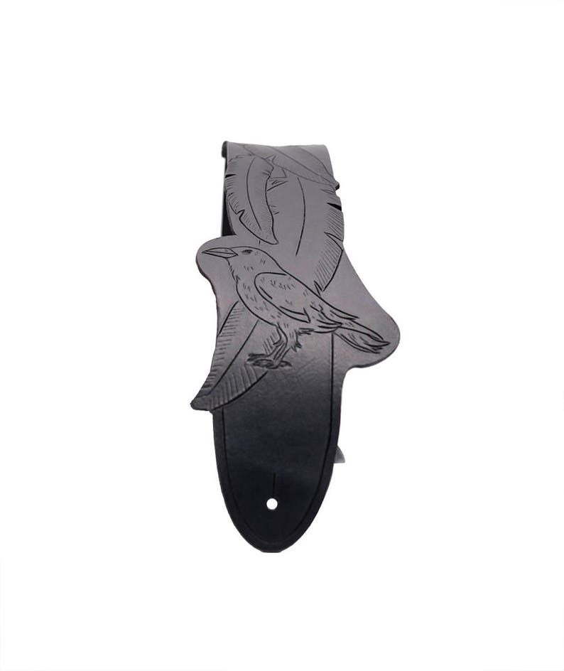 May include: A black guitar strap with a detailed raven and feather design. The strap features a gradient effect, transitioning from light gray to black. A single hole is visible at the bottom for attachment.