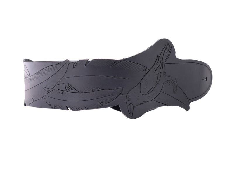 May include: A black leather belt with a decorative feather and bird design. The belt features carved feathers along the strap and a bird motif on the buckle. The belt is a dark gray color and appears to be made of leather.