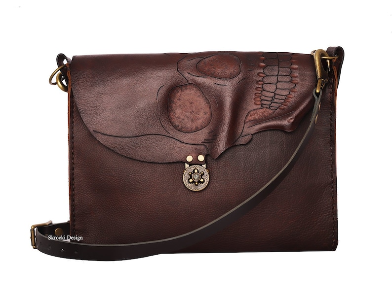 Pode incluir: Bolsa transversal de couro castanho escuro com um design de caveira na aba. A bolsa tem uma al&ccedil;a longa ajust&aacute;vel, um fecho de metal e um detalhe decorativo de metal. A bolsa &eacute; retangular e parece ser feita &agrave; m&atilde;o.