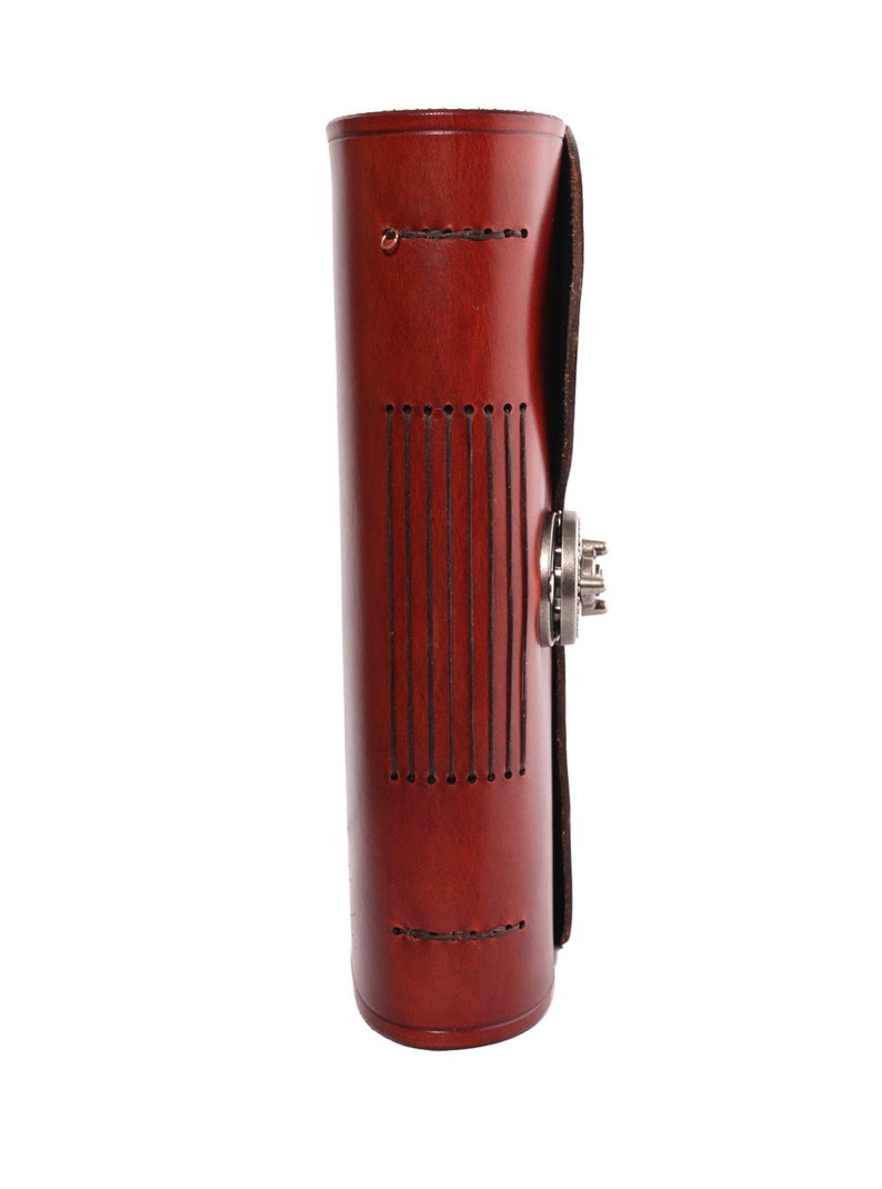 May include: A cylindrical, reddish-brown leather journal with a metal clasp. The journal has vertical stitched lines on the front and a flap closure. The leather has a polished finish, and the journal appears to be new.