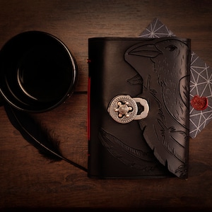 May include: A black leather journal with a raven design, a silver clasp, and red stitching. A black mug, a feather, and a geometric patterned card with a red wax seal are also present on a wooden surface.
