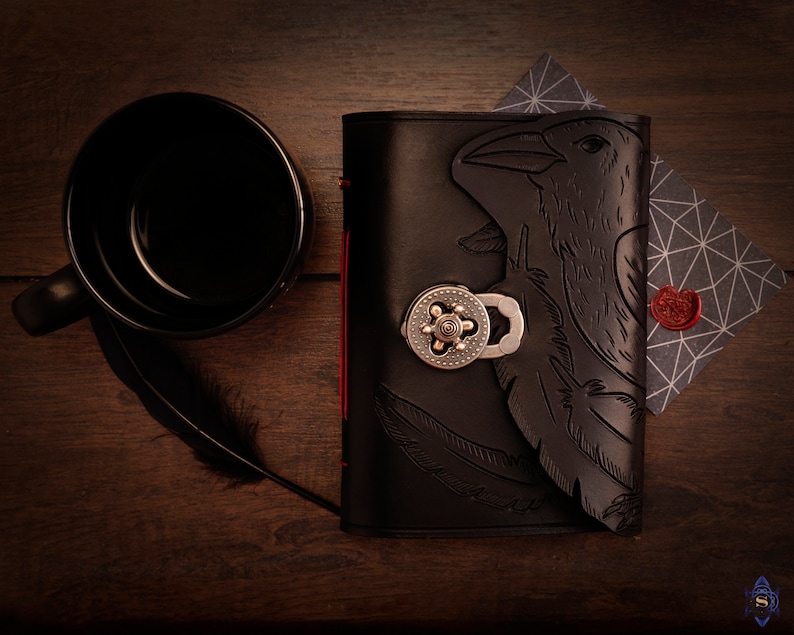 May include: A black leather journal with a raven design, a silver clasp, and red stitching. A black mug, a feather, and a geometric patterned card with a red wax seal are also present on a wooden surface.