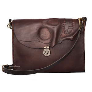 Pode incluir: Bolsa transversal de couro marrom escuro com um design de caveira na aba. A bolsa apresenta uma longa al&ccedil;a ajust&aacute;vel, um fecho decorativo e o texto "SKROCKI DESIGN". A bolsa &eacute; feita de couro texturizado.