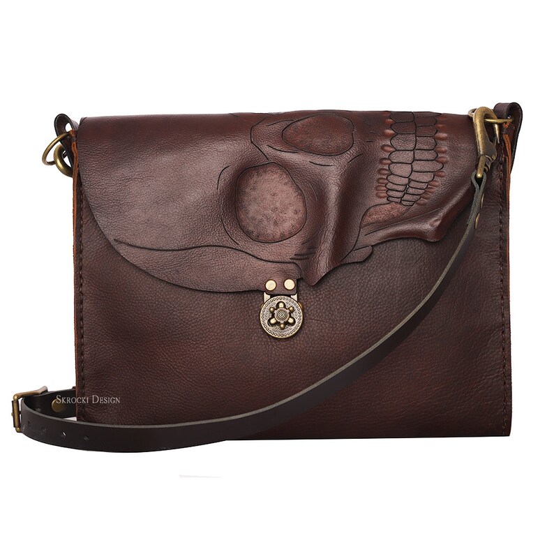 Pode incluir: Bolsa transversal de couro marrom escuro com um design de caveira na aba. A bolsa apresenta uma longa al&ccedil;a ajust&aacute;vel, um fecho decorativo e o texto "SKROCKI DESIGN". A bolsa &eacute; feita de couro texturizado.