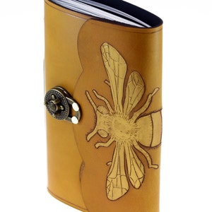 May include: A tan leather journal with a decorative bee design. The journal has a metal clasp and a curved spine. The bee is embossed on the front cover. The pages are visible at the top.