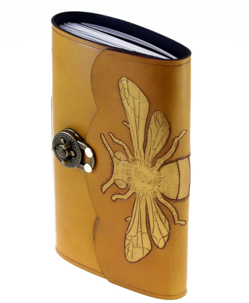 May include: A tan leather journal with a decorative bee design. The journal has a metal clasp and a curved spine. The bee is embossed on the front cover. The pages are visible at the top.