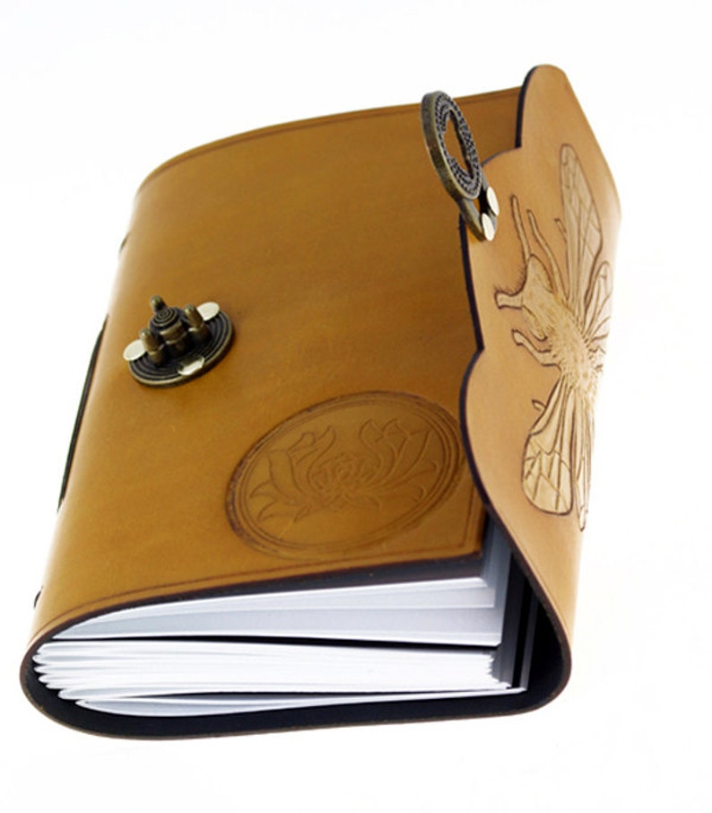 May include: A tan leather journal with a decorative bee and floral design. The journal has a metal clasp and a decorative metal ring. The pages are white and the edges are black.