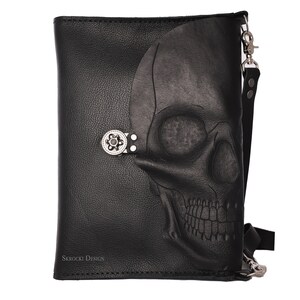 Pode incluir: Bolsa transversal de couro preto com um design de caveira em relevo na frente. A bolsa apresenta um fecho prateado e uma al&ccedil;a longa e ajust&aacute;vel. O texto "Skrocki Design" &eacute; vis&iacute;vel na parte inferior esquerda.