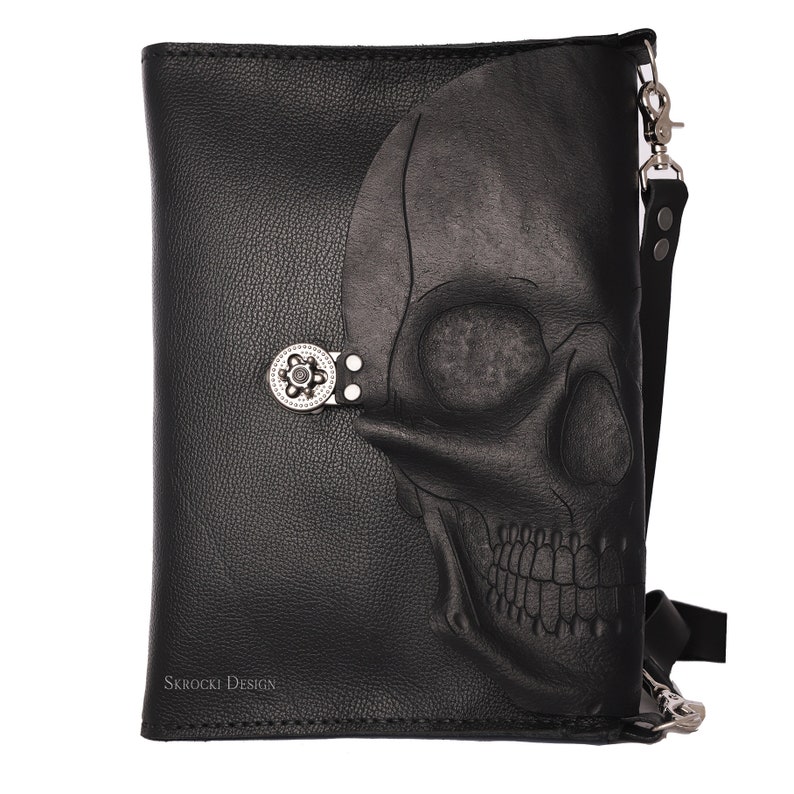 Pode incluir: Bolsa transversal de couro preto com um design de caveira em relevo na frente. A bolsa apresenta um fecho prateado e uma al&ccedil;a longa e ajust&aacute;vel. O texto "Skrocki Design" &eacute; vis&iacute;vel na parte inferior esquerda.