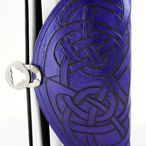 May include: A purple leather journal with a Celtic knot design embossed on the cover. The journal has a silver clasp and a black spine. The pages are white and the journal is open.