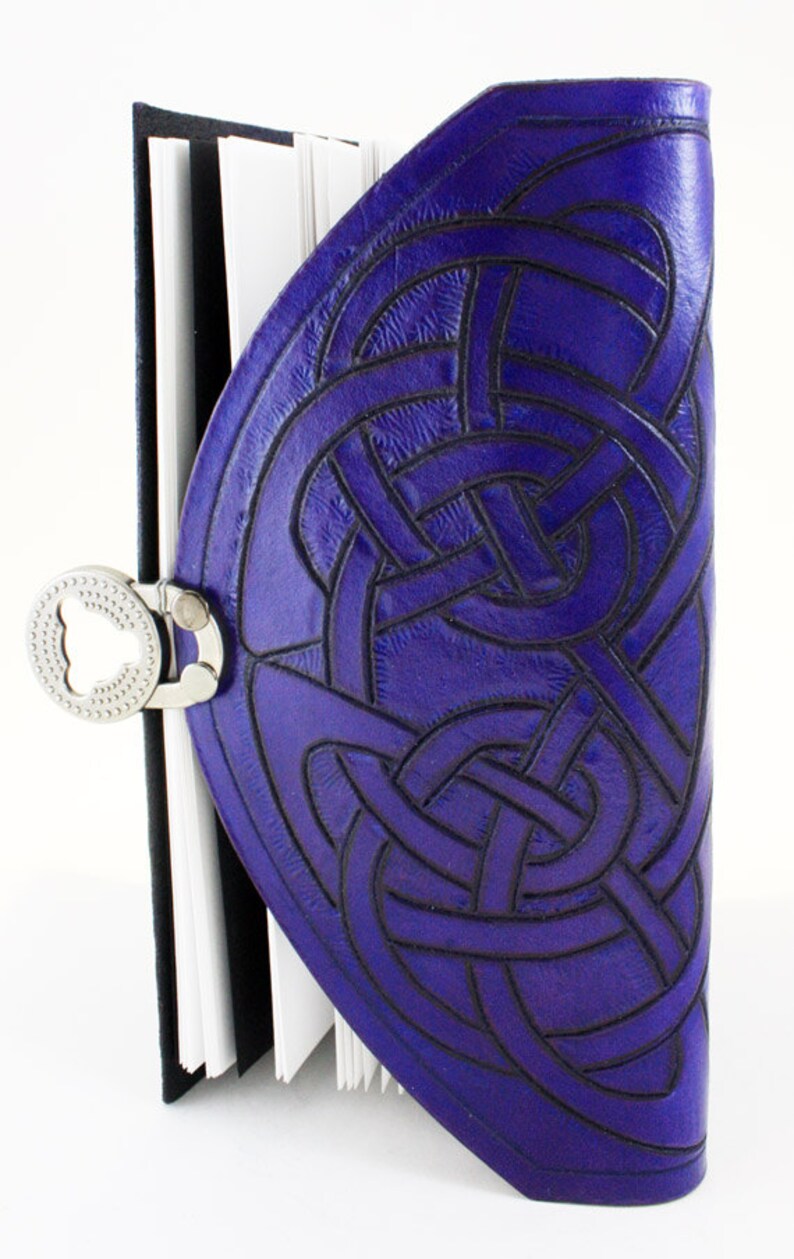 May include: A purple leather journal with a Celtic knot design embossed on the cover. The journal has a silver clasp and a black spine. The pages are white and the journal is open.