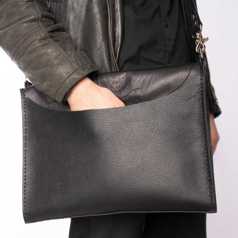 Pode incluir: Uma bolsa mensageiro de couro preto com uma superf&iacute;cie texturizada. A bolsa tem um grande bolso frontal e uma al&ccedil;a de ombro com um fecho prateado. A bolsa &eacute; segurada por uma pessoa que usa uma jaqueta preta.