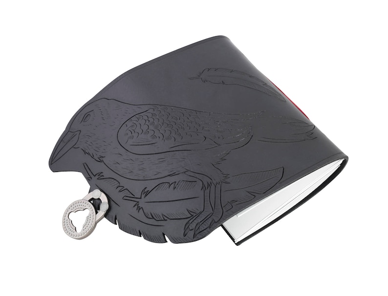 May include: A dark gray journal with a raven design embossed on the cover. The raven is surrounded by feathers. The journal has a silver-colored clasp and a red edge. The pages are white.