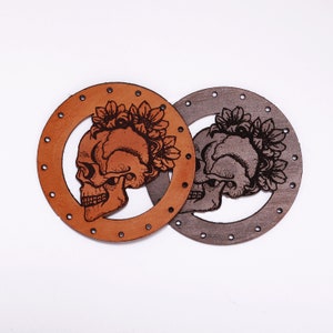 May include: Two leather circles featuring skull designs. One is tan, the other grey. Each has a skull wearing a floral crown in the centre, surrounded by a ring of small holes. The skulls are detailed with shading and line work.