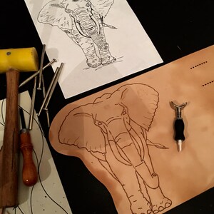 May include: An elephant outline is drawn on a piece of paper and on a piece of tan leather. A yellow mallet, nails, and other leatherworking tools are arranged around the elephant drawings. The image suggests a leathercraft project.