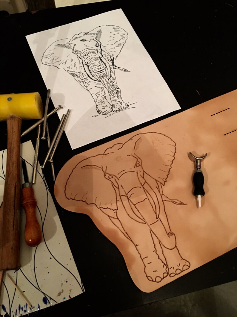 May include: An elephant outline is drawn on a piece of paper and on a piece of tan leather. A yellow mallet, nails, and other leatherworking tools are arranged around the elephant drawings. The image suggests a leathercraft project.