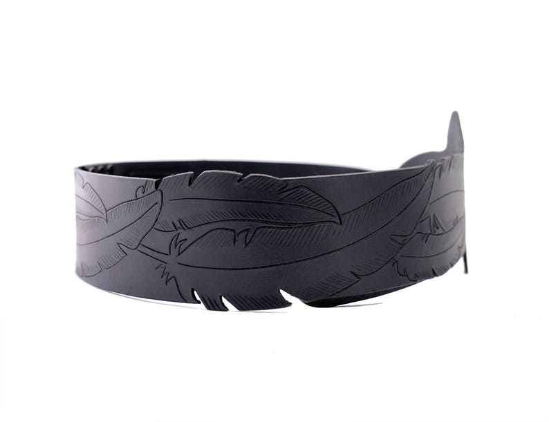 May include: A black, feather-detailed headband. The headband is made of a dark material and features an embossed feather design. The design wraps around the entire band, creating a decorative accessory.