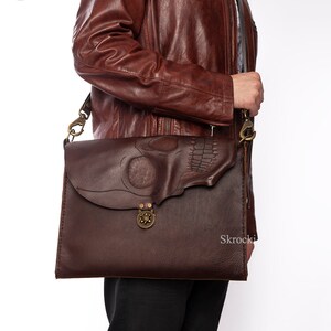Pode incluir: Bolsa mensageiro de couro castanho escuro com um design de caveira na aba. A bolsa apresenta um fecho e ferragens de lat&atilde;o, e uma longa al&ccedil;a ajust&aacute;vel. A bolsa tem aproximadamente 30 cm de largura e 25 cm de altura.