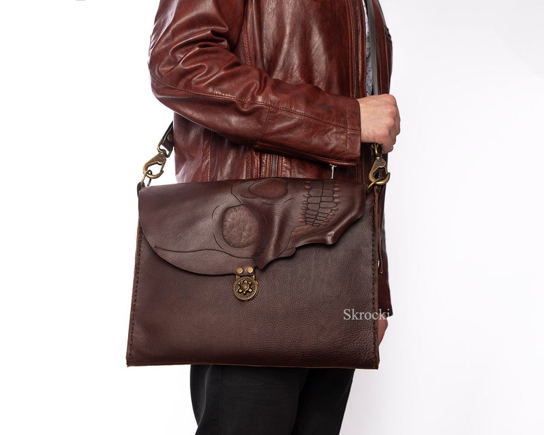Pode incluir: Bolsa mensageiro de couro castanho escuro com um design de caveira na aba. A bolsa apresenta um fecho e ferragens de lat&atilde;o, e uma longa al&ccedil;a ajust&aacute;vel. A bolsa tem aproximadamente 30 cm de largura e 25 cm de altura.