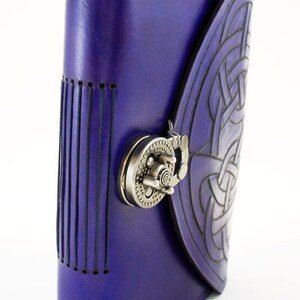 May include: A purple leather journal with a Celtic knot design embossed on the cover. The journal features a metal clasp closure and stitched detailing along the spine and edges. The cover is a deep shade of purple.