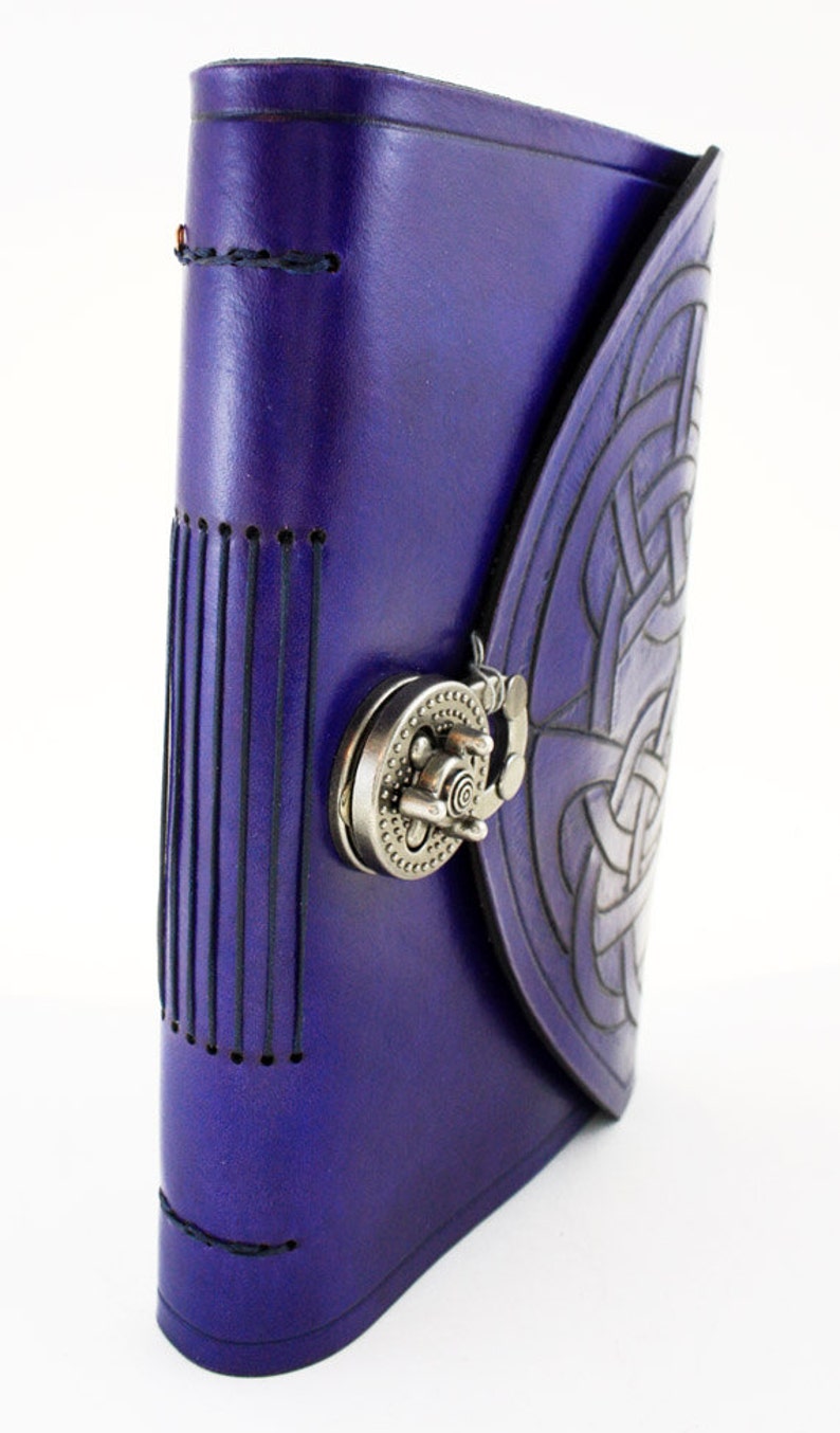 May include: A purple leather journal with a Celtic knot design embossed on the cover. The journal features a metal clasp closure and stitched detailing along the spine and edges. The cover is a deep shade of purple.