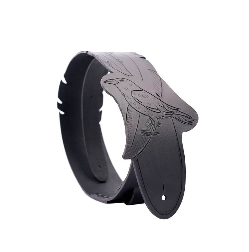 May include: A black leather guitar strap featuring a detailed raven design. The strap has a curved shape with a raven illustration on the shoulder area and a hole for adjustment. The strap is designed to be worn over the shoulder.