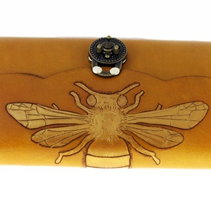May include: A tan leather wallet with a detailed embossed bee design. The wallet has a decorative clasp and a scalloped edge. The bee is centered on the front of the wallet, with intricate wing and body details.