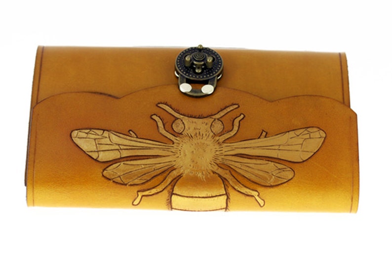 May include: A tan leather wallet with a detailed embossed bee design. The wallet has a decorative clasp and a scalloped edge. The bee is centered on the front of the wallet, with intricate wing and body details.
