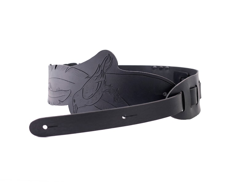 May include: A black guitar strap with a detailed, embossed design featuring a bird. The strap is made of leather and has adjustable slots for length. The design is centered on the shoulder area of the strap.