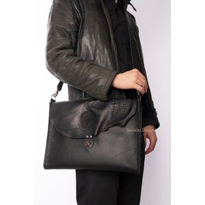 Pode incluir: Bolsa mensageiro de couro preto com um design de caveira em relevo na aba. A bolsa tem uma al&ccedil;a de ombro e um fecho de metal. A bolsa retangular tem costuras vis&iacute;veis. O texto "SKROCKI DESIGN" &eacute; vis&iacute;vel.