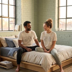 Puede incluir: Dos personas con camisetas blanquecinas a juego con un dibujo lineal, sentadas en una cama con ropa de cama de color claro. La habitación tiene una pared de ladrillo verde claro y grandes ventanas.